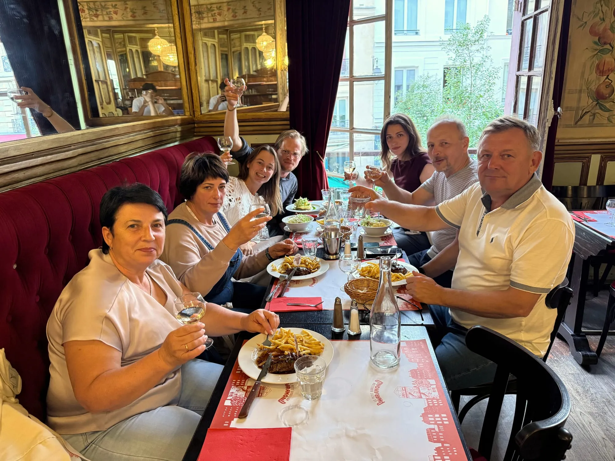 Seven guests raise wine glasses over steak frites at a classic Parisian bistro table